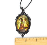 A gothic-style oval pendant necklace with a black ornate frame and a stained-glass window design featuring a witch's hat, spiderwebs, and a bat. A wooden ruler is placed below the pendant to show its scale against a white background.
