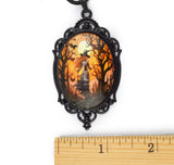 A Halloween-themed oval pendant with a witch silhouette, bats, and bare trees in orange and black, set in a black ornate frame. A wooden ruler is placed below the pendant to show scale against a white background.