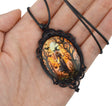 A gothic-style oval pendant necklace with a black ornate metal frame and a dark cord, held in a person's hand. The pendant features a colorful printed illustration of a ghostly figure in a spooky forest at night under a full moon.