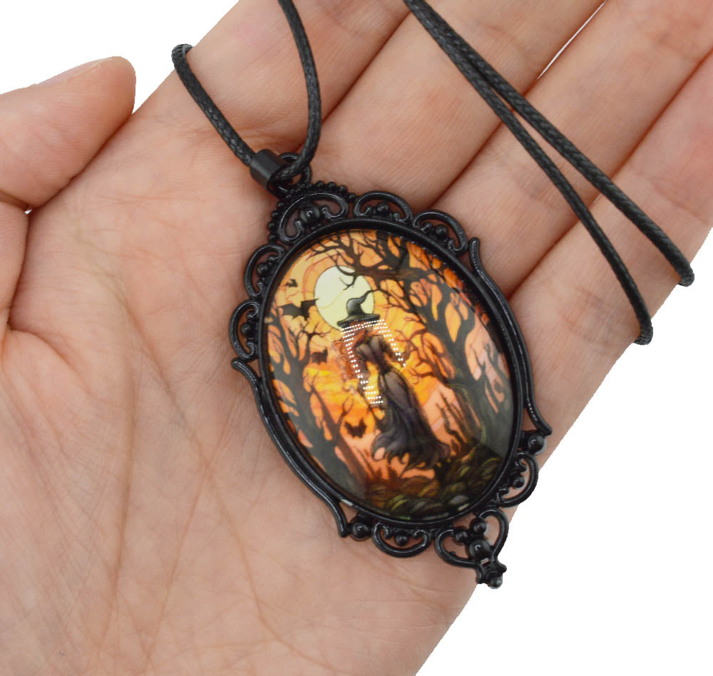 A gothic-style oval pendant necklace with a black ornate metal frame and a dark cord, held in a person's hand. The pendant features a colorful printed illustration of a ghostly figure in a spooky forest at night under a full moon.