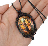 A gothic-style oval pendant necklace with a black ornate metal frame and a dark cord, held in a person's hand. The pendant features a colorful printed illustration of a ghostly figure in a spooky forest at night under a full moon.