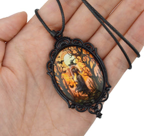 A gothic-style oval pendant necklace with a black ornate metal frame and a dark cord, held in a person's hand. The pendant features a colorful printed illustration of a ghostly figure in a spooky forest at night under a full moon.