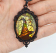 A hand holding an oval pendant with a black ornate frame and a stained-glass-style illustration in yellow, green, brown, and orange tones.