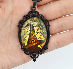 A hand holding an oval pendant with a black ornate frame and a stained-glass-style illustration in yellow, green, brown, and orange tones.