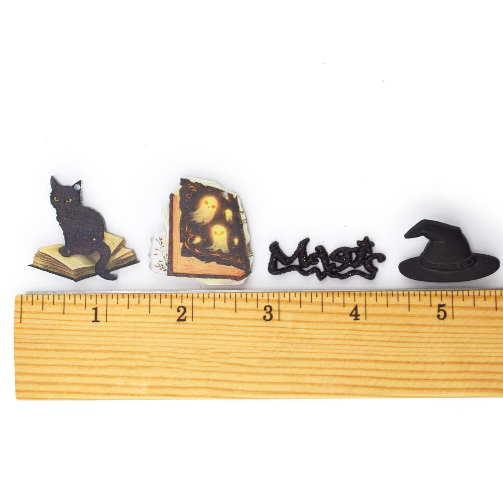 Four small Halloween-themed decorative items arranged above a wooden ruler on a white background: a black cat sitting on an open book, a haunted book with glowing yellow faces, a black spider-shaped ornament, and a black witch hat.