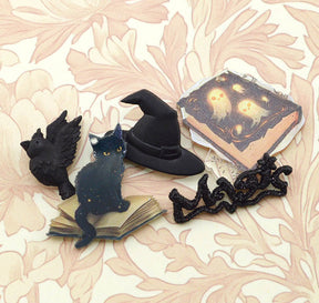 A set of Halloween-themed decorative items arranged on a beige floral patterned surface, including a black witch hat, a black cat figurine on a miniature book, a black raven figurine, a black spider decoration, and a small gothic book with glowing yellow ghost illustrations.