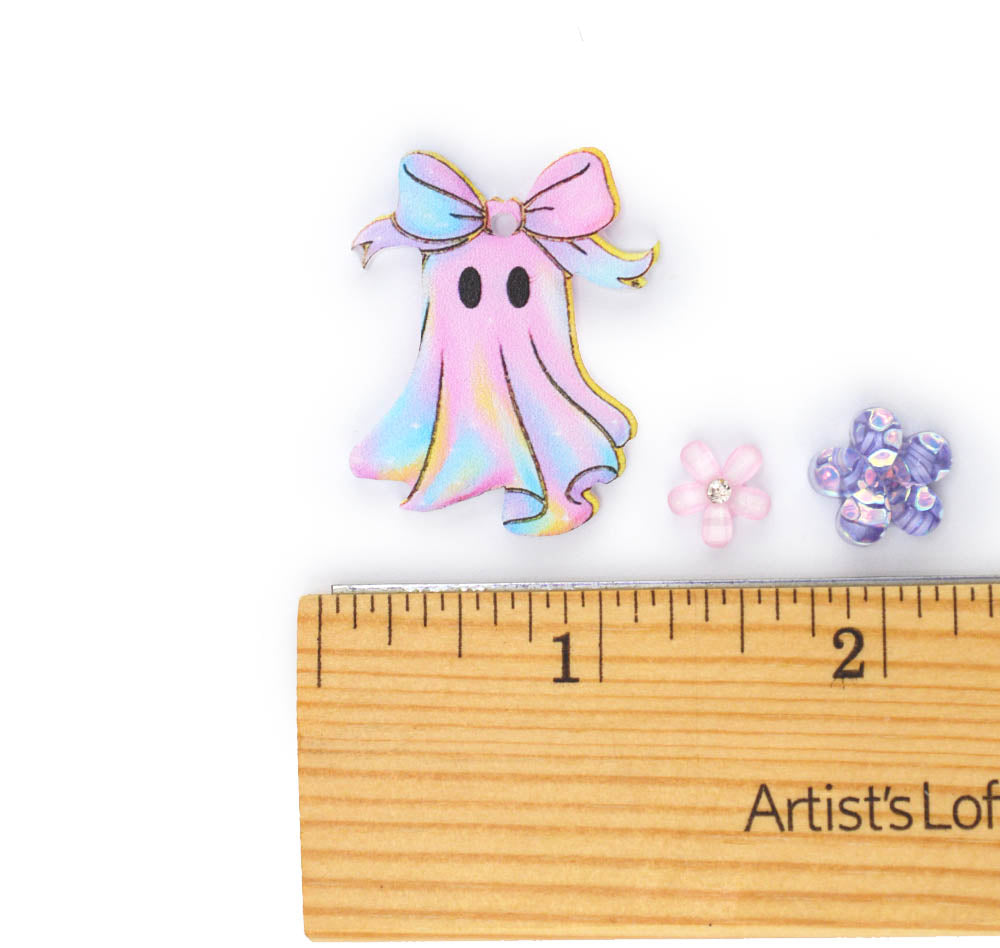 An iridescent pastel ghost-shaped embellishment with a bow, shown next to a wooden ruler and two small flower beads on a white background.