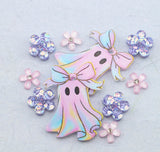 A pair of pastel ghost charms with bows, surrounded by pink and iridescent flower embellishments on a textured light blue background.