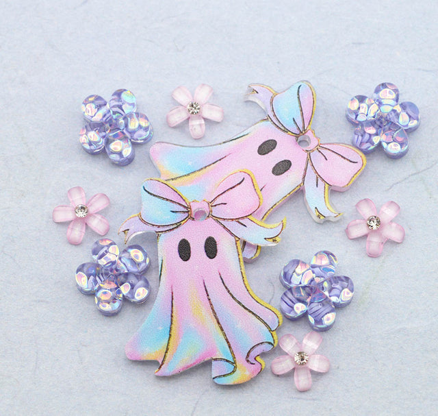 A pair of pastel ghost charms with bows, surrounded by pink and iridescent flower embellishments on a textured light blue background.