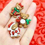 Three shank-back Christmas bear buttons under a Christmas tree with a Santa costume, a present, and a joy sign